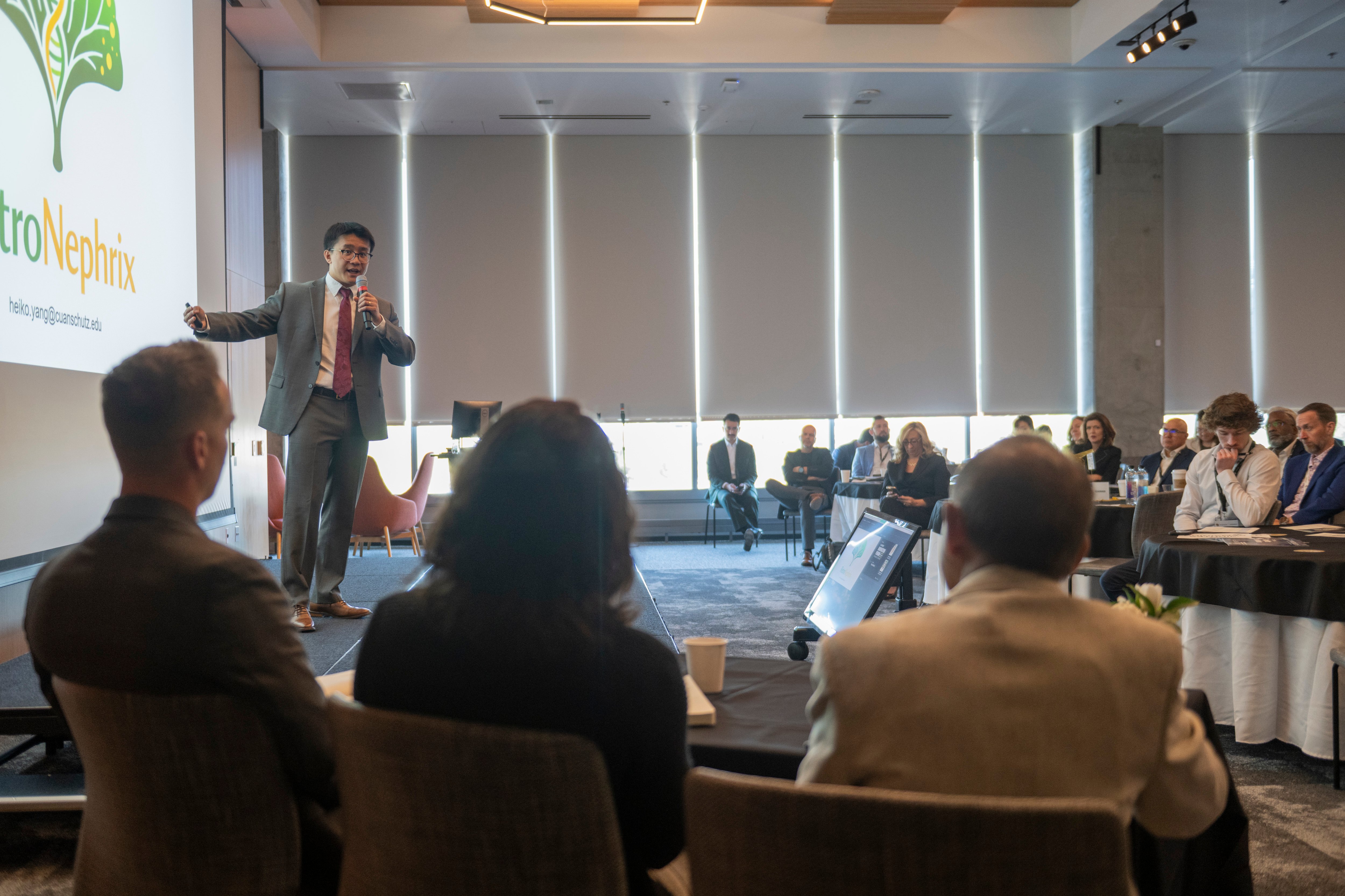 Heiko Yang points towards to a screen with his pitchdeck, microphone in hand, with a crowd in front of him. The camera is placed behind the three members of the event panel's shoulders.