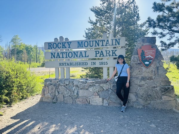 A woman smiles beside a sign for the Rocky Mountain National Park.