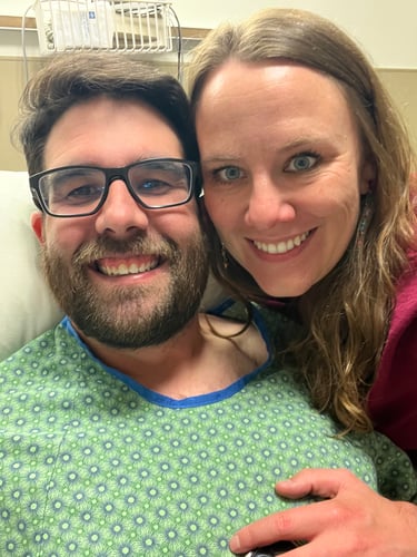 A man, dressed in a hospital gown, and a woman smile for a selfie.