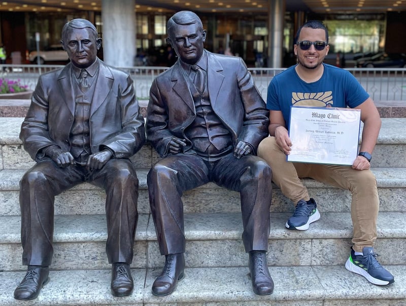 A man sitting on stairs to the far right of two outdoor statues of men that are also sitting on stairs. 