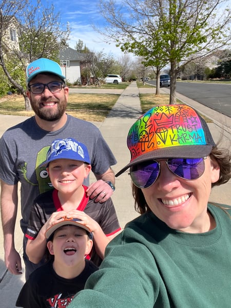 Kristie Click, wearing a colorful hat, smiles while taking a selfie with her husband, Bobby Click, and two sons as they walk outside on a sunny day.