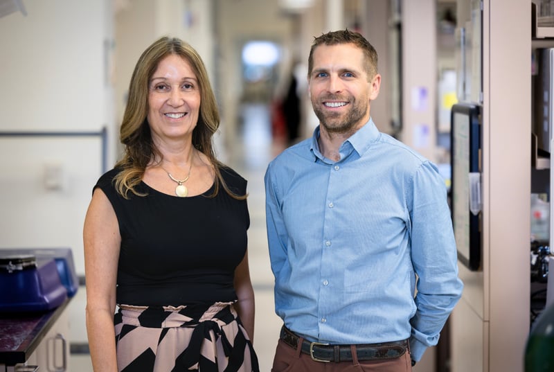 Two researchers, a woman wearing a black top and a man wearing a long-sleeved blue dress shirt, smile while standing in a building at the CU Anschutz campus.