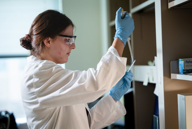 A female scientist, wearing a white coat, blue gloves, and protective eyewear, works in a lab.