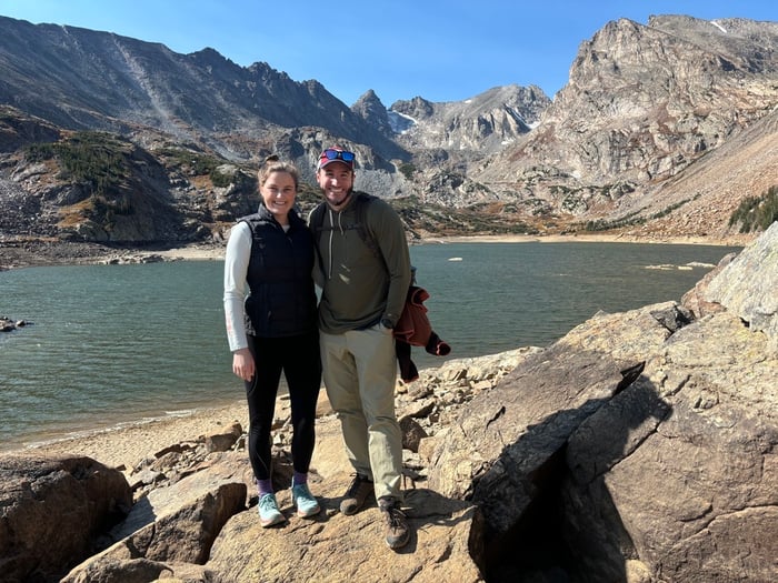 Two people smiling while standing in front of a lake near mountains.