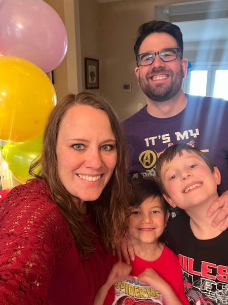 Kristie Click smiles while taking a selfie with her husband, Bobby Click, and two sons while inside a house, standing beside some colorful, inflated balloons.