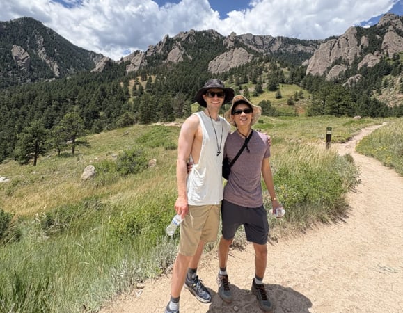 Two people smile and pose for a photo on a sunny day while hiking on a trail near the mountains.