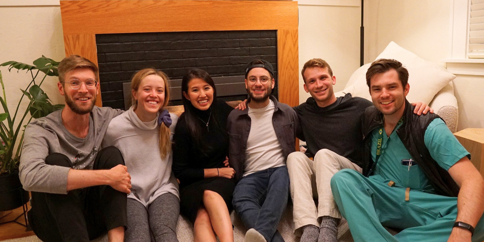 A group of six adults smile together while sitting on the floor near a fireplace.