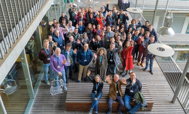 A large group of researchers smile and wave for a photo.