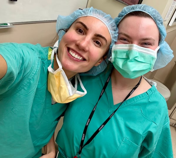 Two health care workers wearing scrubs smile for a selfie.