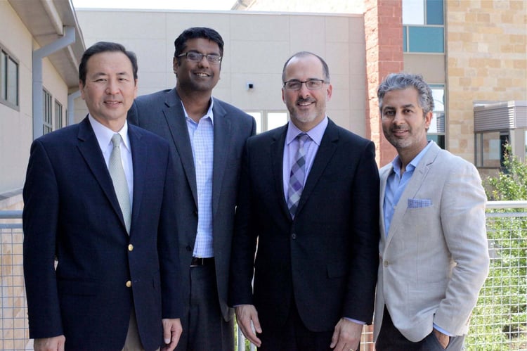 Four ophthalmologists, dressed in suits, stand and smile for a group photo outside of a building.