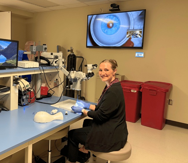 A woman smiles beside a microscope. She is holding tools to operate on a synthetic eyeball.
