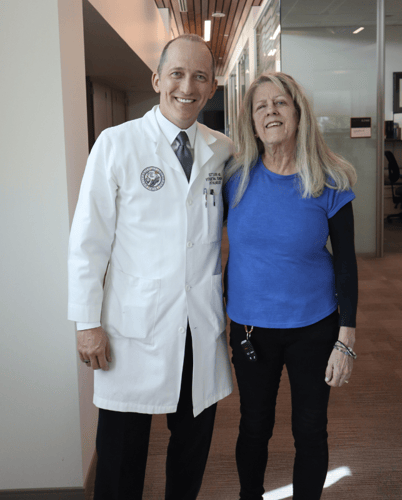 At CU Anschutz, a male doctor wearing a white coat smiles beside a woman wearing a blue shirt with black sleeves.