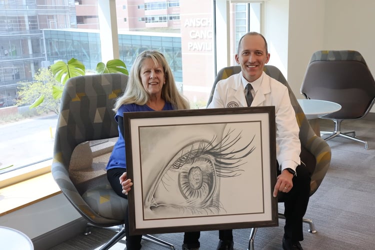 Diane Ciampa and Scott Oliver smile while sitting in chairs. They are each holding up one side of a framed art piece of an eyeball.