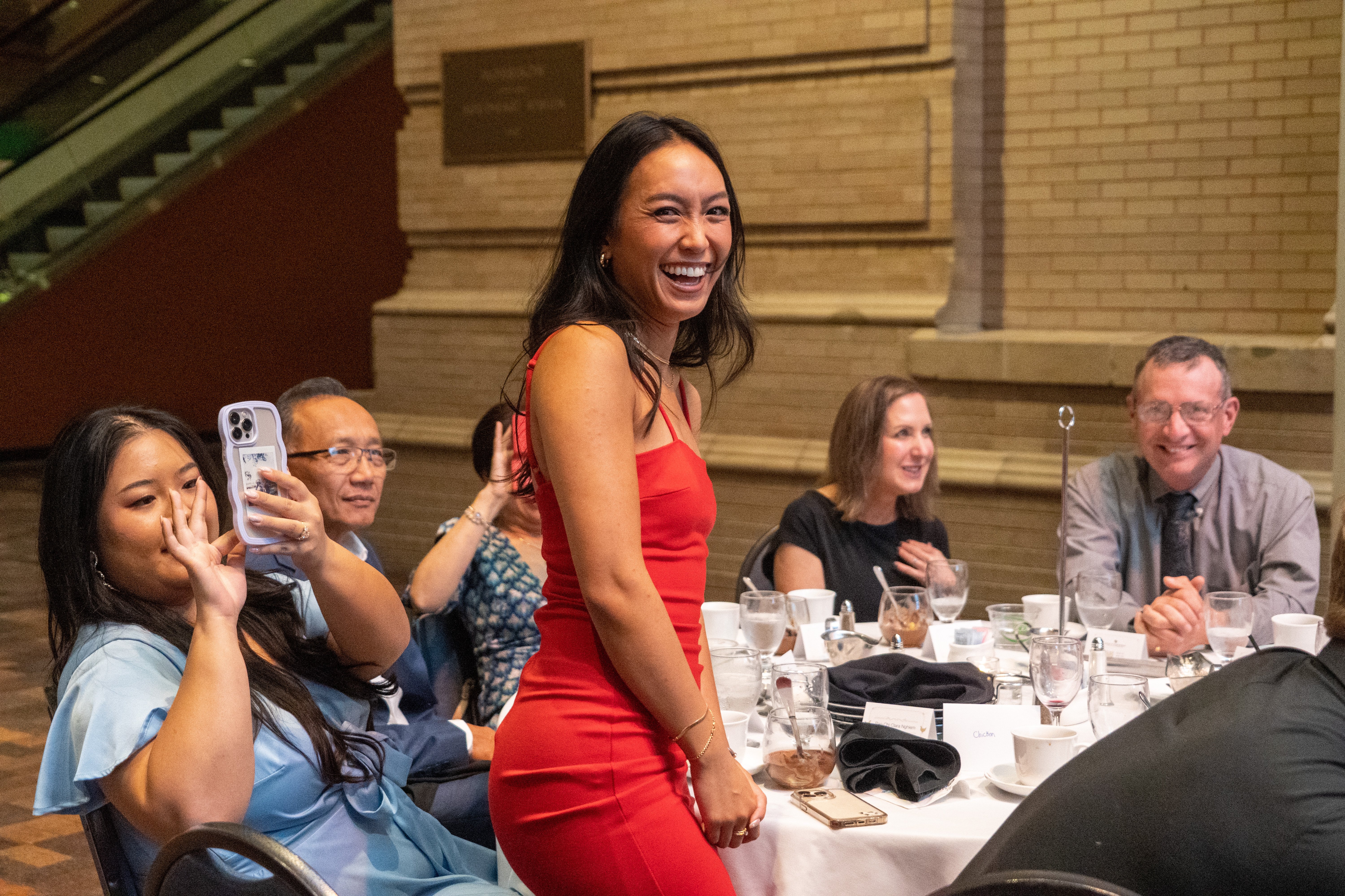 woman smiling, standing at table surrounded by family and friends
