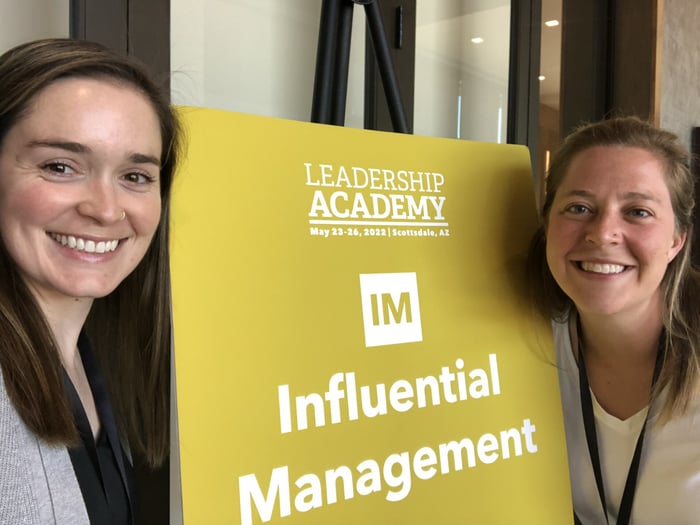 Two physician assistants smile beside a poster that says: "Leadership Academy IM Influential Management."