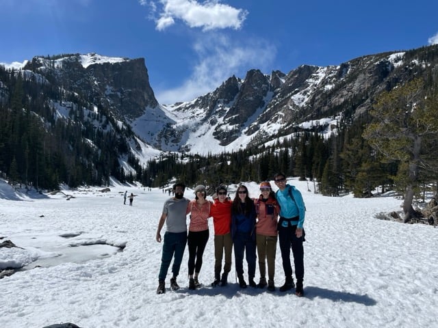 Six adults smile outdoors for a group photo with snowy mountains in the background.