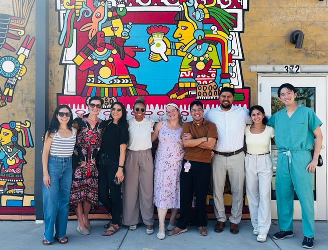 A group of people smile while standing outdoors in front of a mural on a wall.