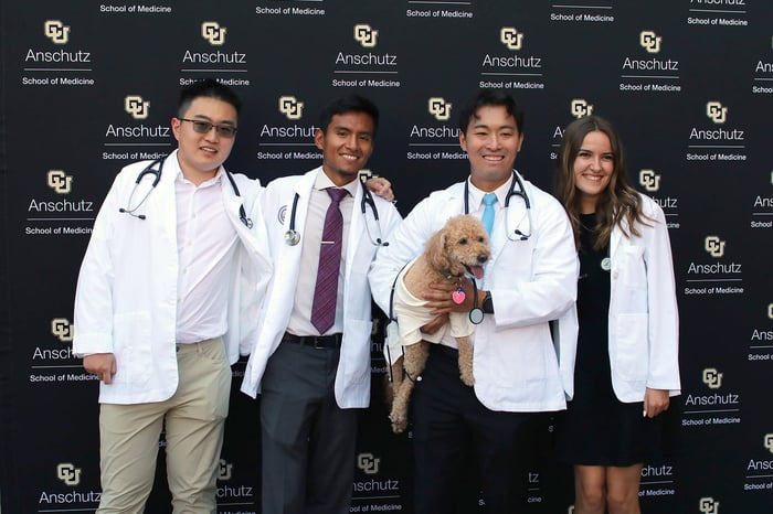 Four students donning their white coats smile in front of a CU Anschutz banner while one student holds a small brown dog.
