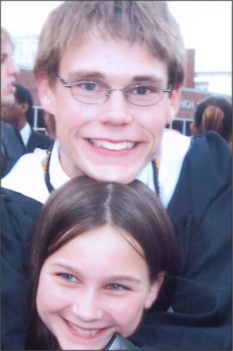 A young man smiles with his younger sister, placing his head above hers for a photo.