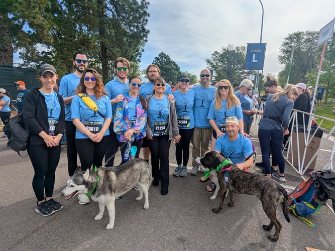 A group of adults dressed in blue smile for a photo while standing outside on a street. There are two dogs standing in front, beside the group.