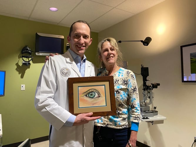 Scott Oliver, MD, and Diane Ciampa smile while in an eye clinic. Oliver is holding up a pastel drawing of an eye that Ciampa made.
