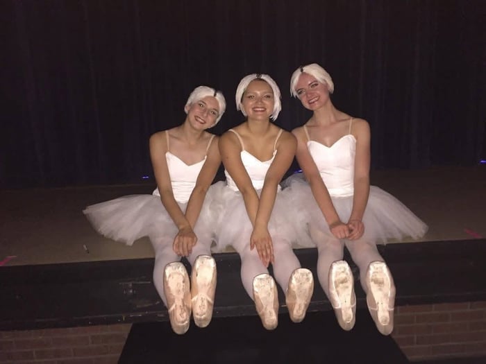Three girls dressed in tutus and wearing blonde wigs smile while sitting on a stage.