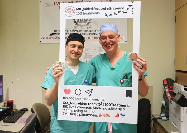 Drew Kern, MD, MS, and Steven Ojemann, MD, smile in their scrubs while holding up cookies that have their faces on them. They are standing within a cardboard frame that recognizes the 100th high-intensity focused ultrasound case being performed at UCHealth University of Colorado Hospital.