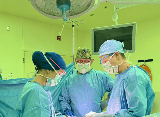 Three people, two men and a woman, in scrubs and caps, in an operating room. They are looking down toward the table.