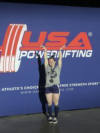 Tessa Petersell poses for a picture in front of the USA Powerlifting logo.