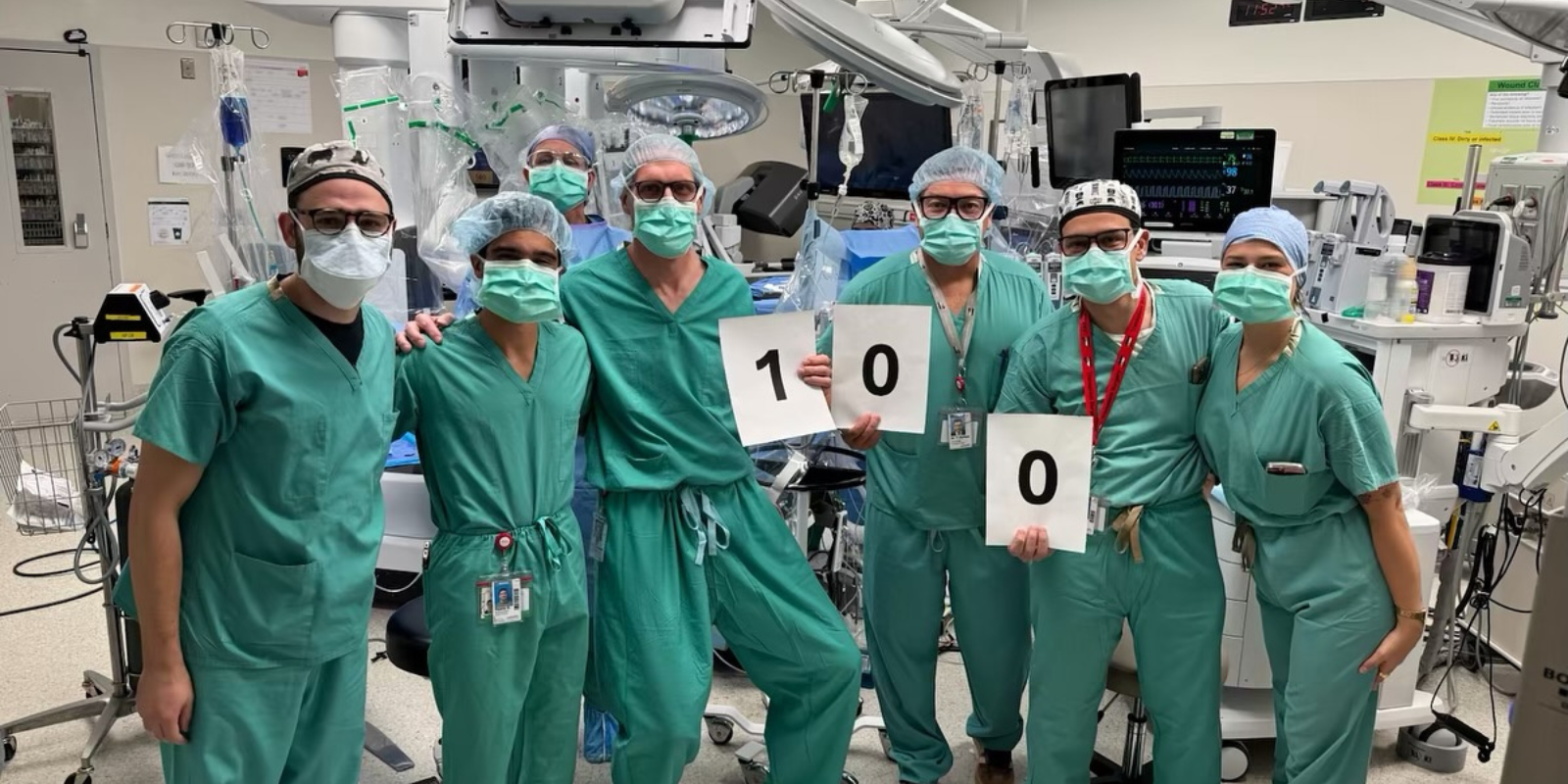 transplant surgery team, in masks and scrubs, celebrating 100th transplant with homemade sign