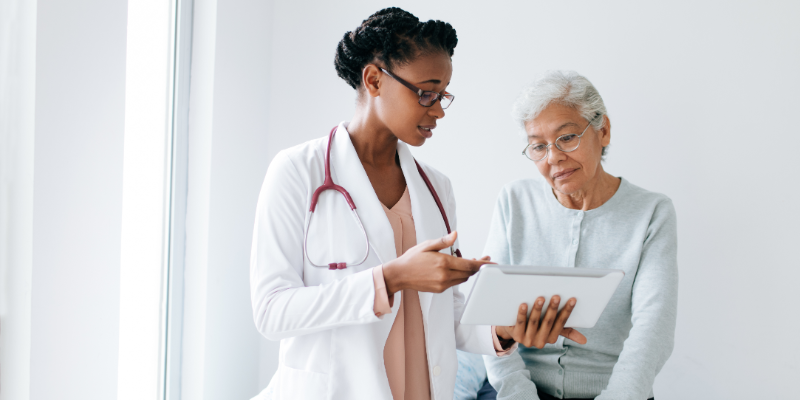 A female doctor wearing a white coat and a stethoscope, clip board in hand, speaks to an elderly seated woman.