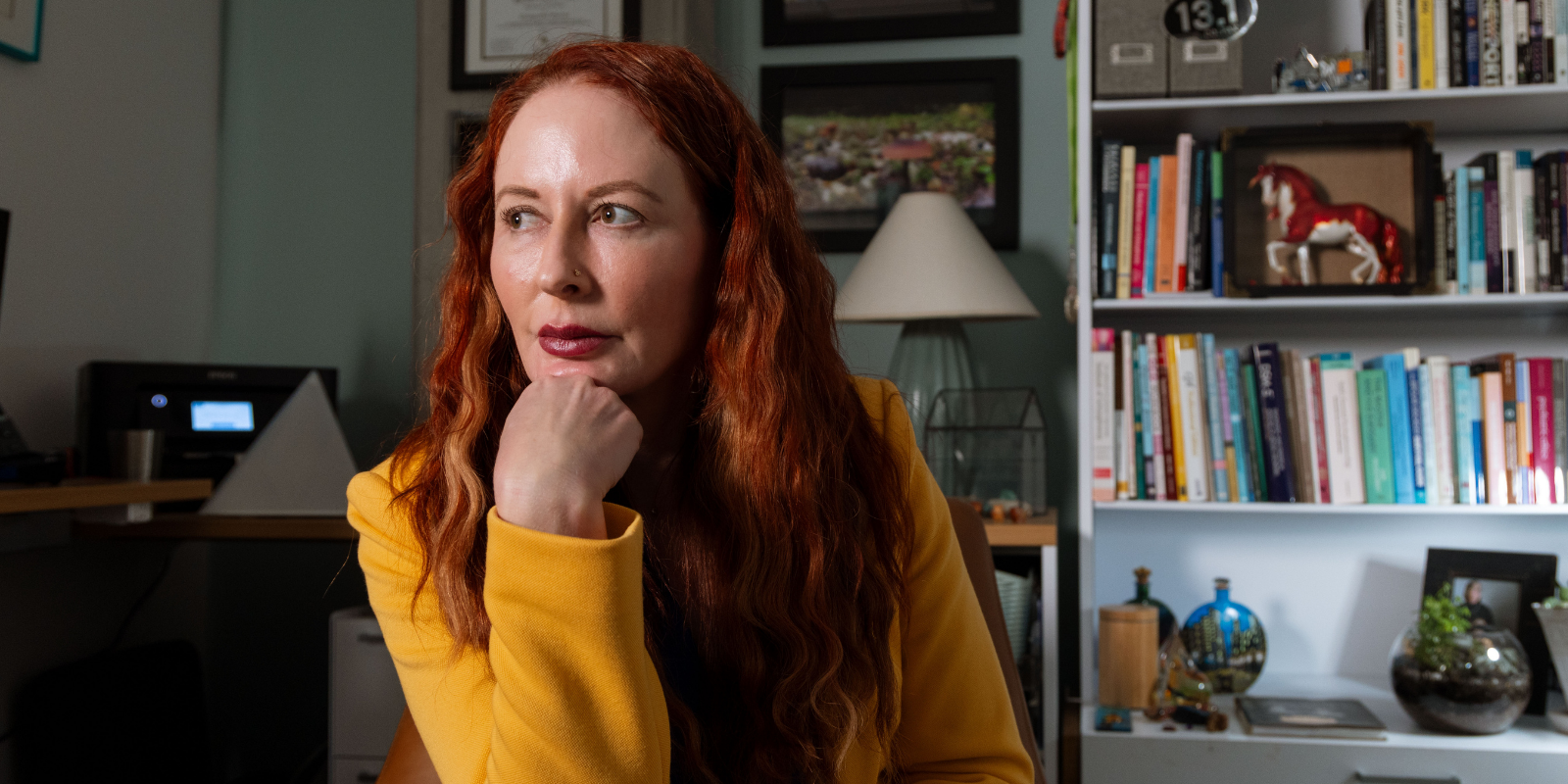 Rachel Davis sits in front of her floor-to-ceiling office bookcase with her chin on her fist and her eyes looking away from the camera in a bright gold blazer
