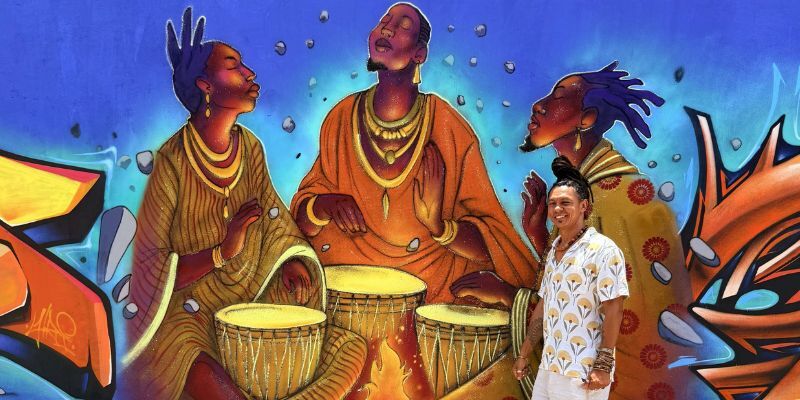 Mural artist Eder Muniz stands in front of his work "The first was the first master" depicting three ancestral figures around a fire with drums.