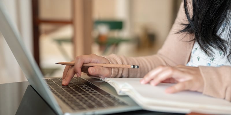 Cropped image of a woman working on her laptop, taking notes in a book and typing on the keyboard.