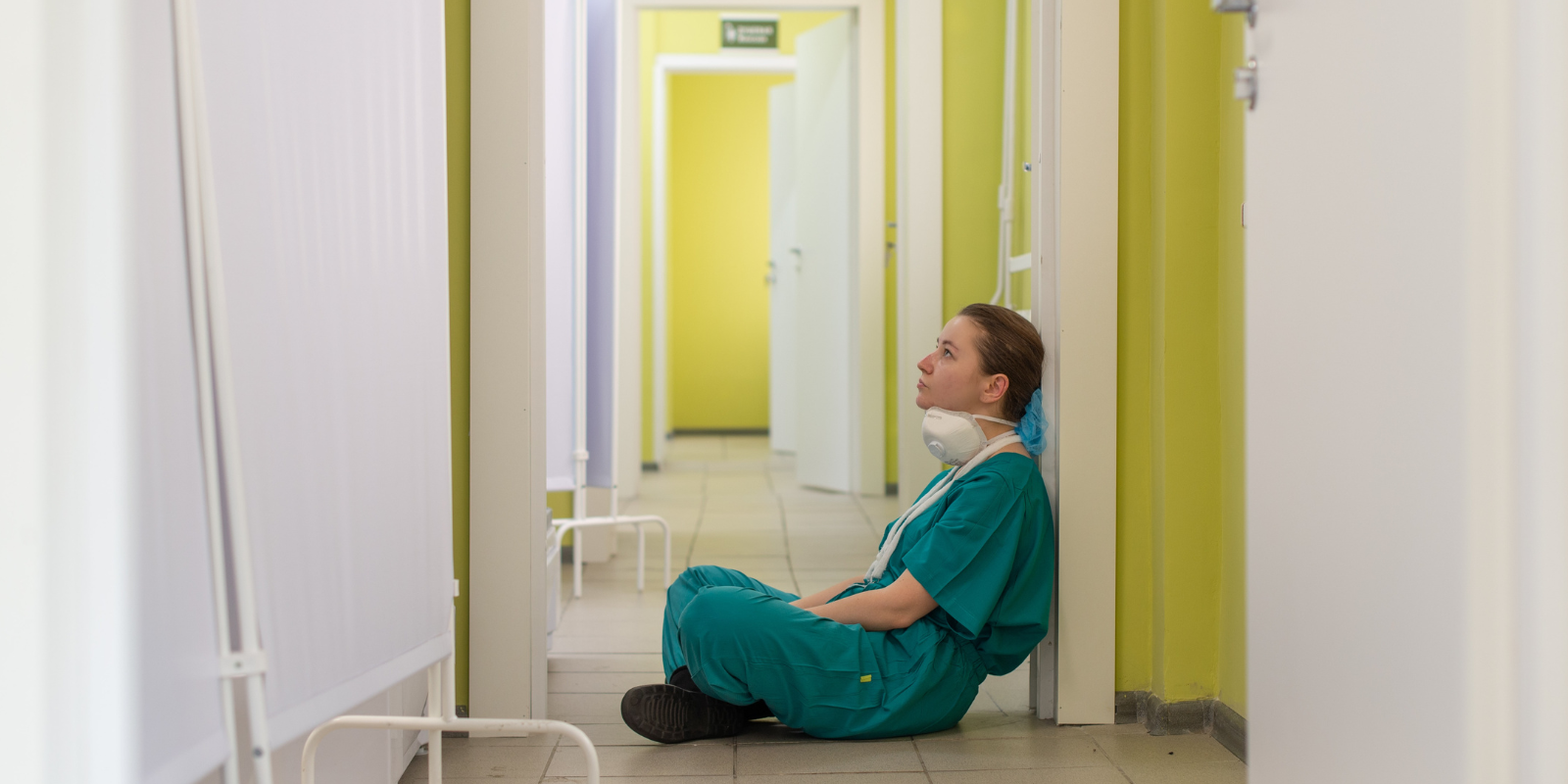 A medical provider sits alone on the floor of a hospital hallway looking upward. 