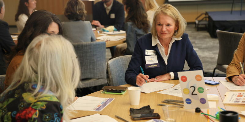 A group of professionals participates in a small-group discussion at a workshop, taking notes around a table marked with a "2" sign.