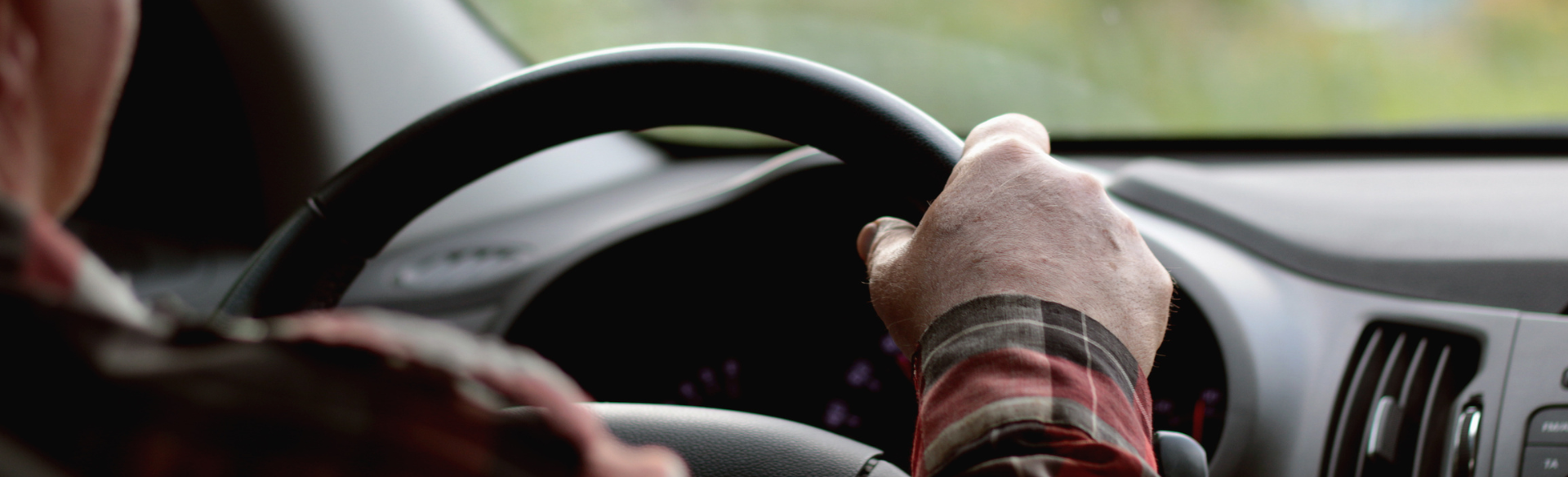 Two older hands grip the steering wheel of a car. 