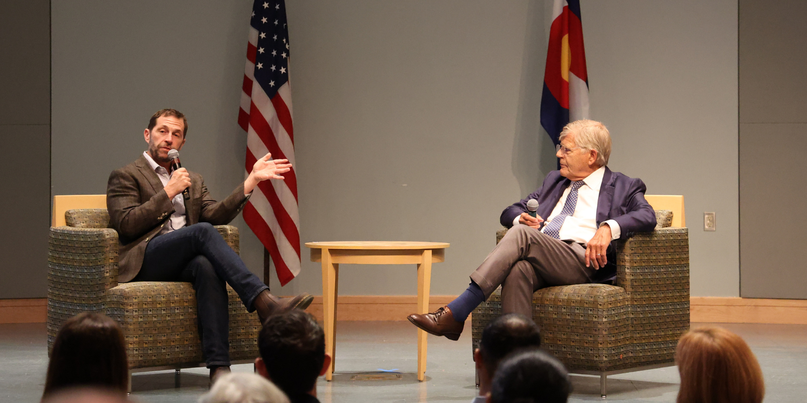 U.S. Rep. Jason Crow and Chancellor Don Elliman on stage, seated, with an American and Colorado flag behind them. 