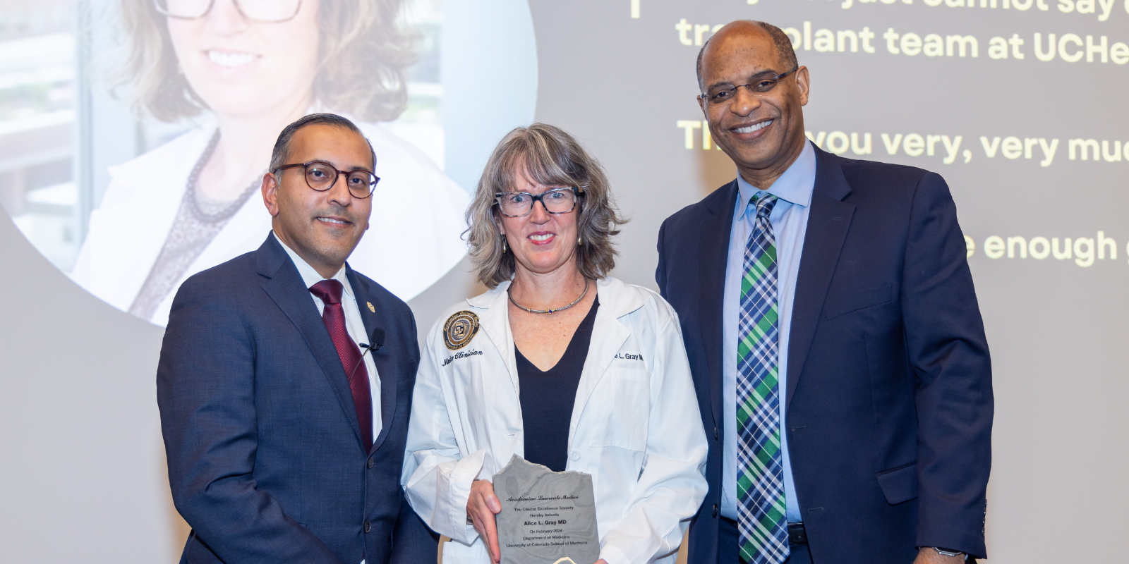 Three people standing and smiling. In the center, a woman holds a plaque.