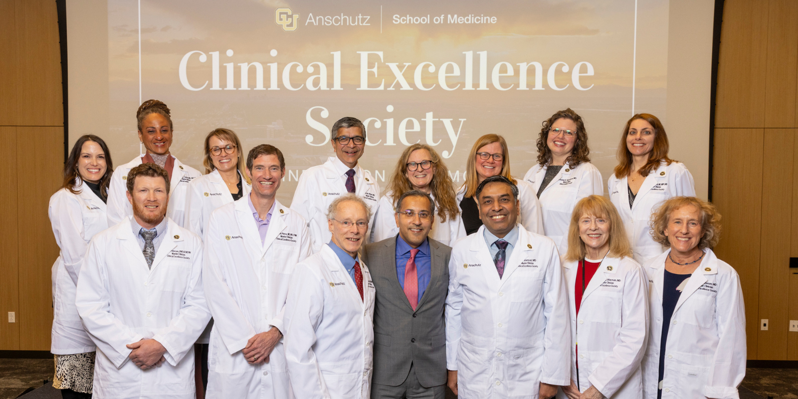 A group of 15 clinicians, most of whom are wearing white coats, smile for a group photo to celebrate the newest Clinical Excellence Society cohort.