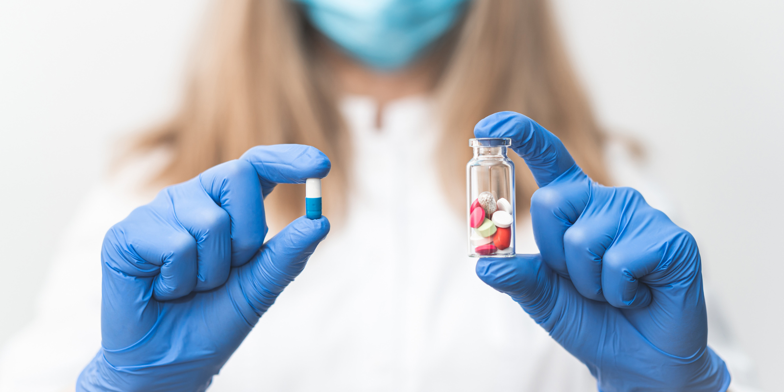 A woman wearing blue gloves holding a pill in one hand and a vial of pills in the other hand.