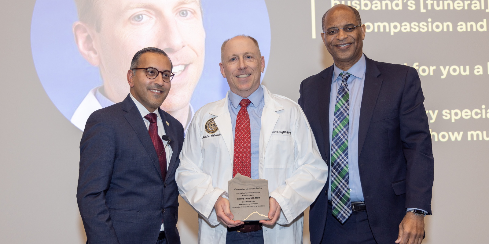 Three people standing beside one another. The man in the center holds an award.