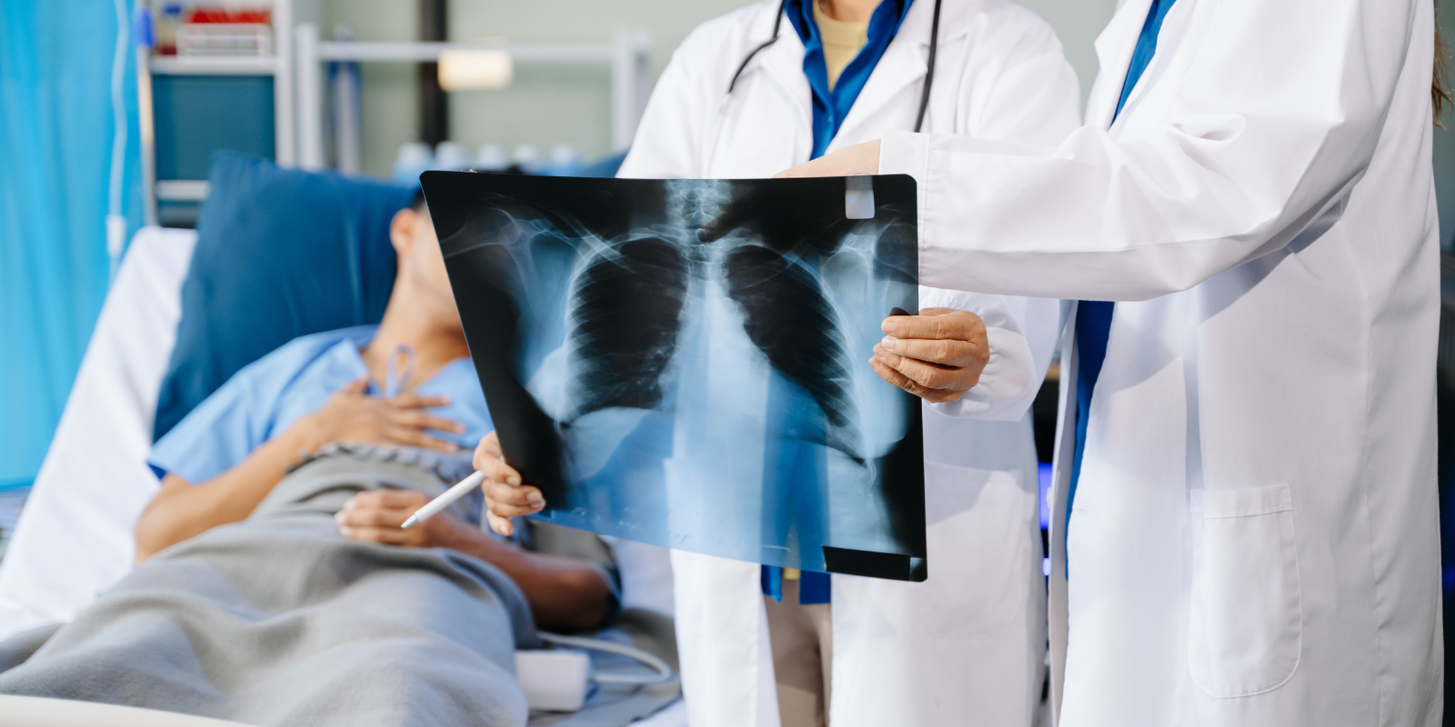 Two doctors look at an MRI scan of a chest while a patient lays in a hospital bed behind them.
