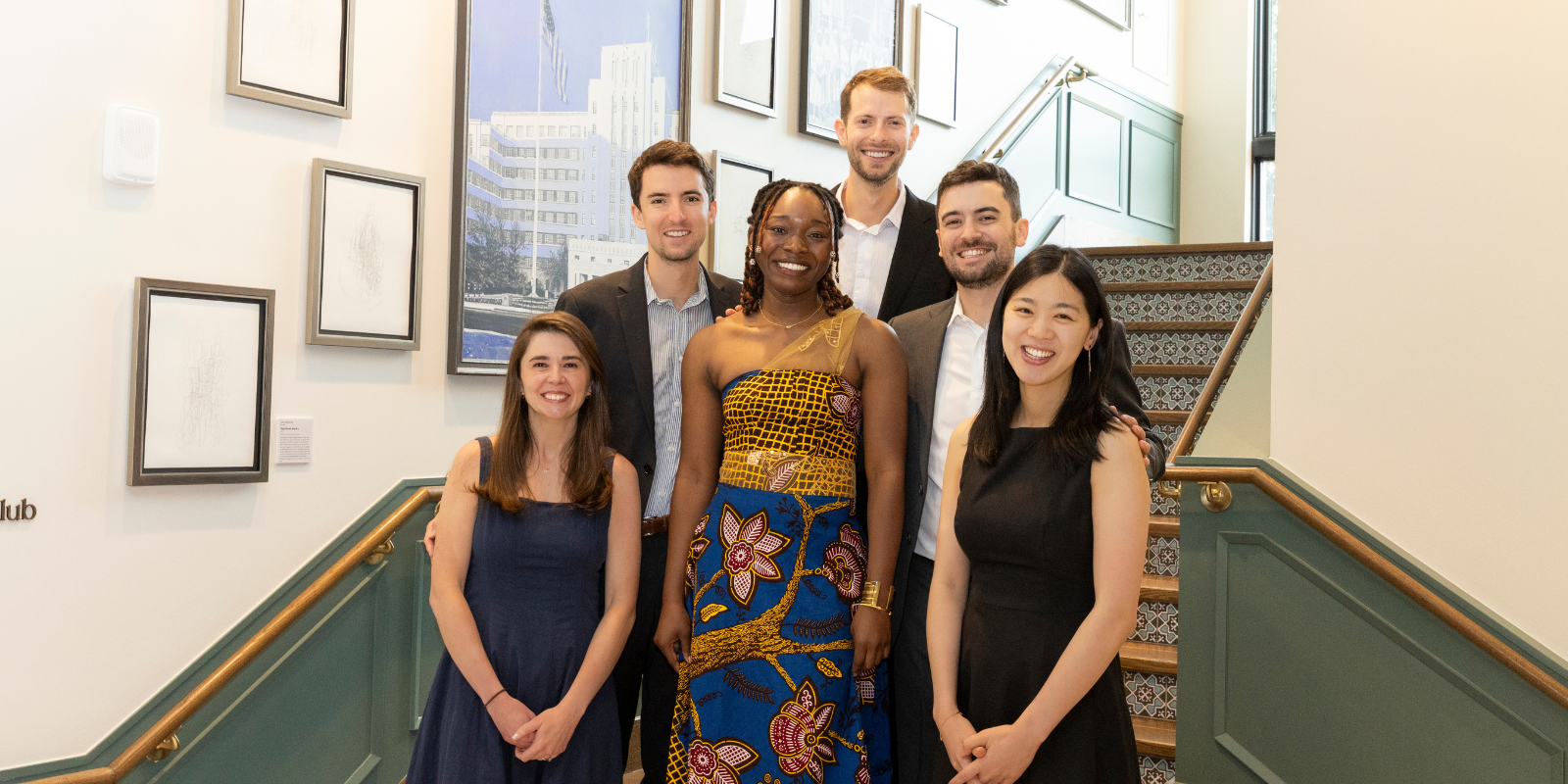 Six adults, three men and three women, smile while standing on a staircase.