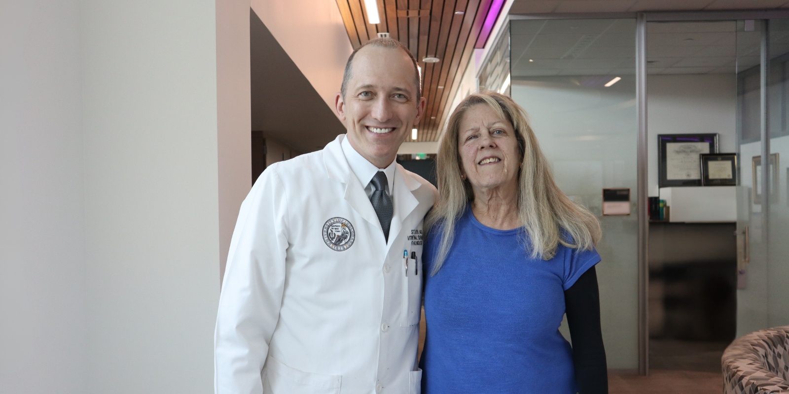 A male doctor wearing a white coat smiles beside a woman wearing a blue shirt with black sleeves.