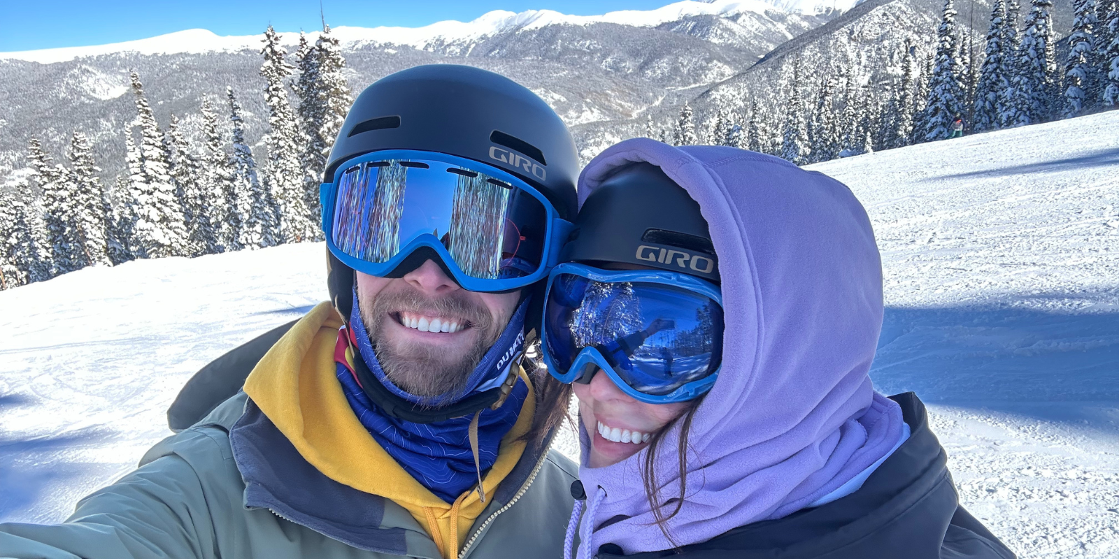 A man and a woman, both wearing ski goggles and helmets, take a selfie while in the snowy mountains.