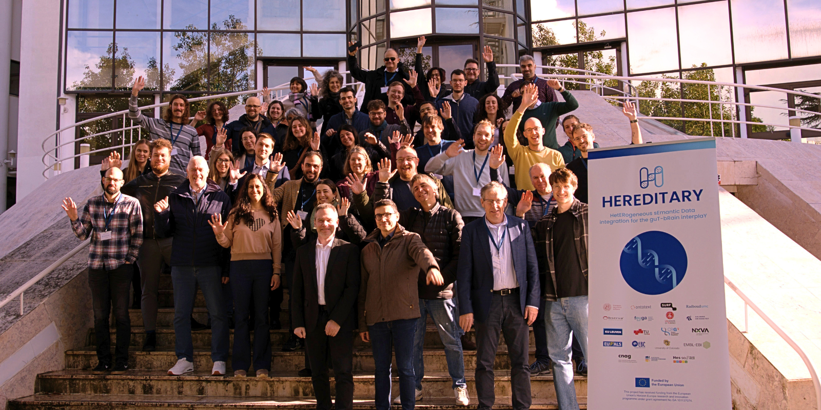 A large group of researchers stand on steps outside of a building, with many smiling and waving for a group photo. To the right, there is a sign for the HEREDITARY project.