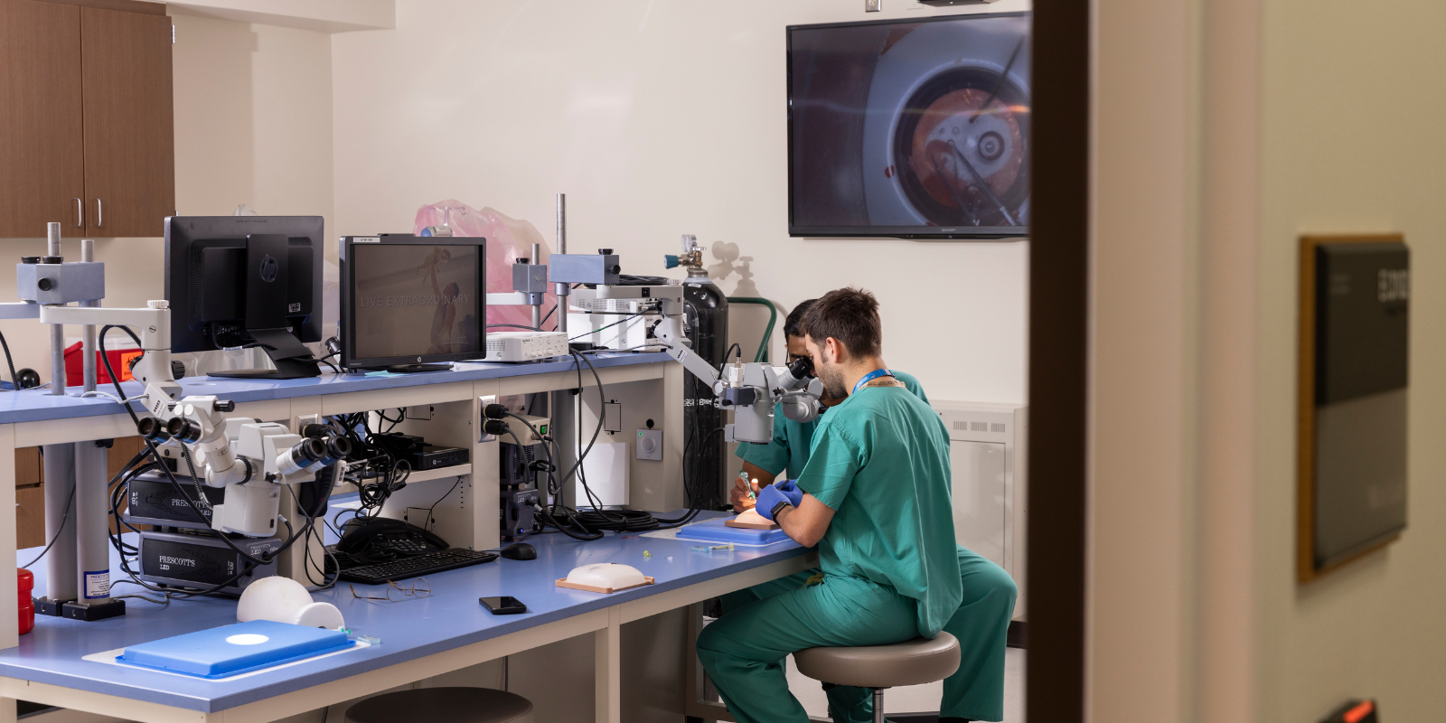 Two men dressed in scrubs look into a microscope in a lab.