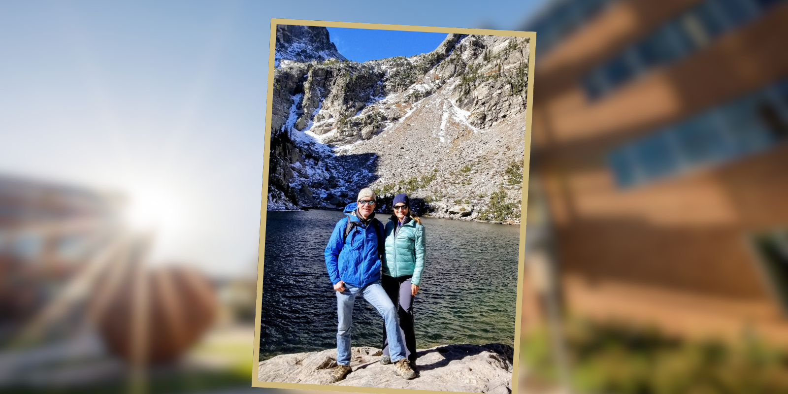 Jim, in a blue coat and jeans, poses for a photo with his wife Anita, in a teal jacket, on a hike near a high altitude lake in the Rocky Mountains.