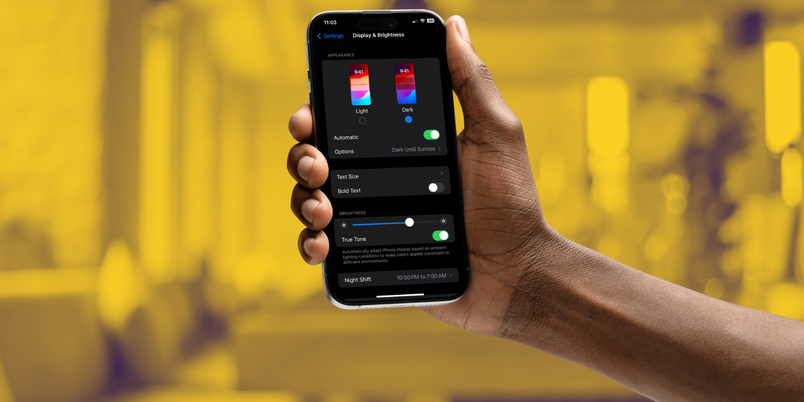 A hand holds out an iPhone, which shows lighting settings. Yellow and purple blurred background. 